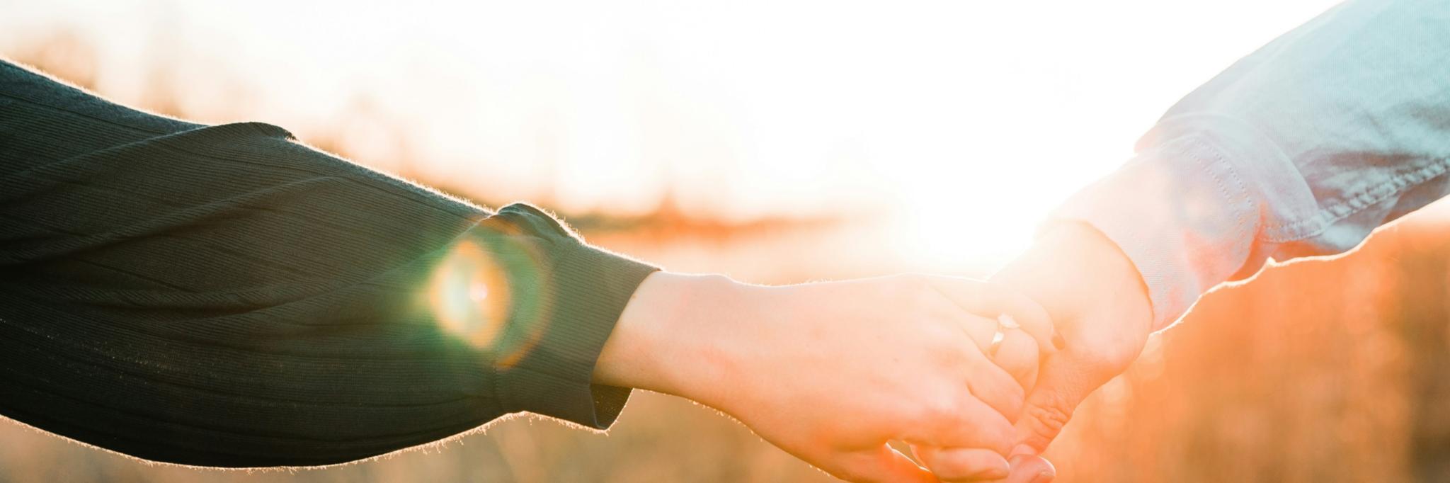 Zwei Hände halten sich fest, im Hintergrund ist eine Wiese und die Sonne geht unter