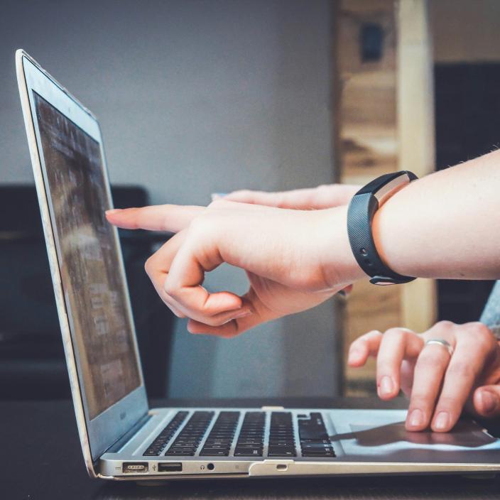 eine Hand zeigt auf eine Stelle an einen Laptop-Bildschirm, an dem jemand arbeitet.