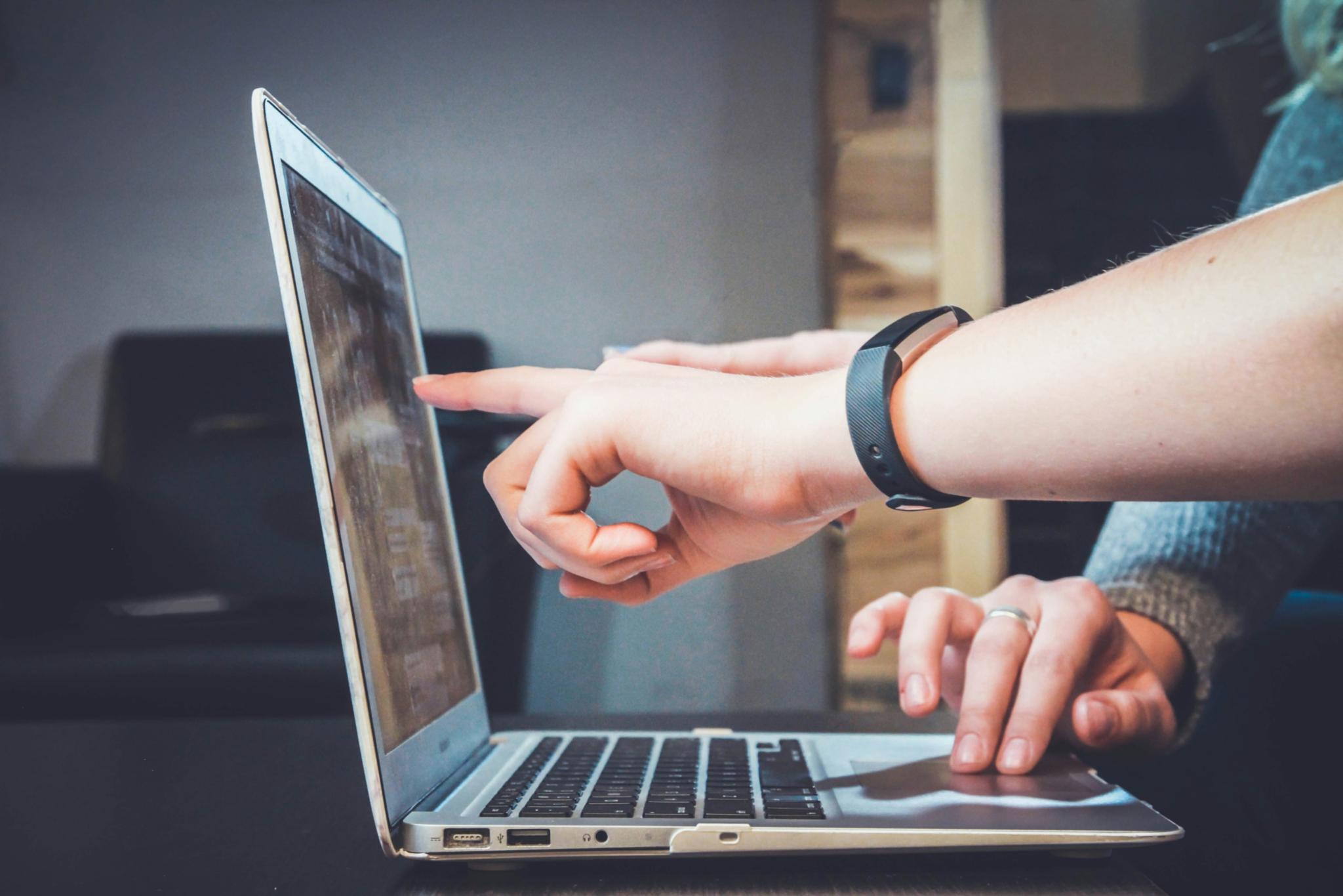 eine Hand zeigt auf eine Stelle an einen Laptop-Bildschirm, an dem jemand arbeitet.