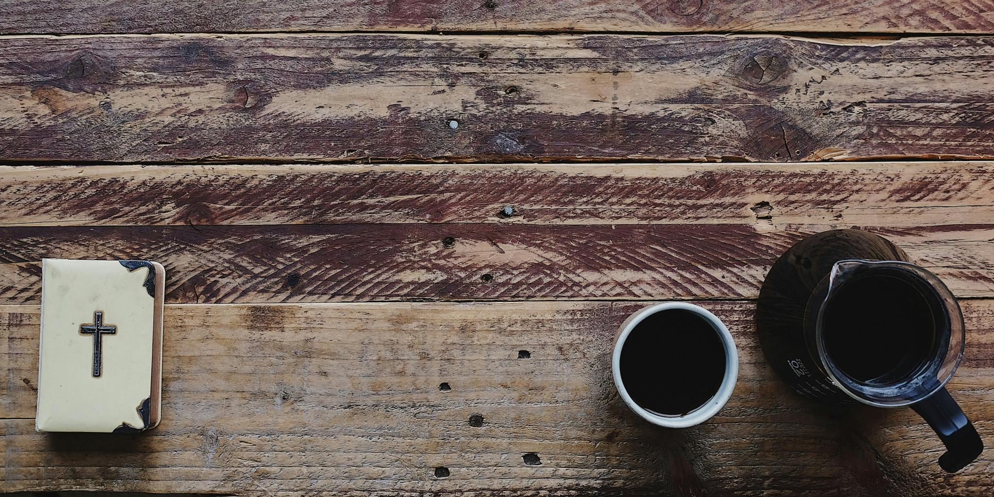 Auf einem alten Holztisch liegt links unten eine weiße Bibel mit einem Kreuz und Verzierungen aus Silber und au der rechten Seite eine Kanne Kaffee, daneben steht ein Becher mit Kaffee