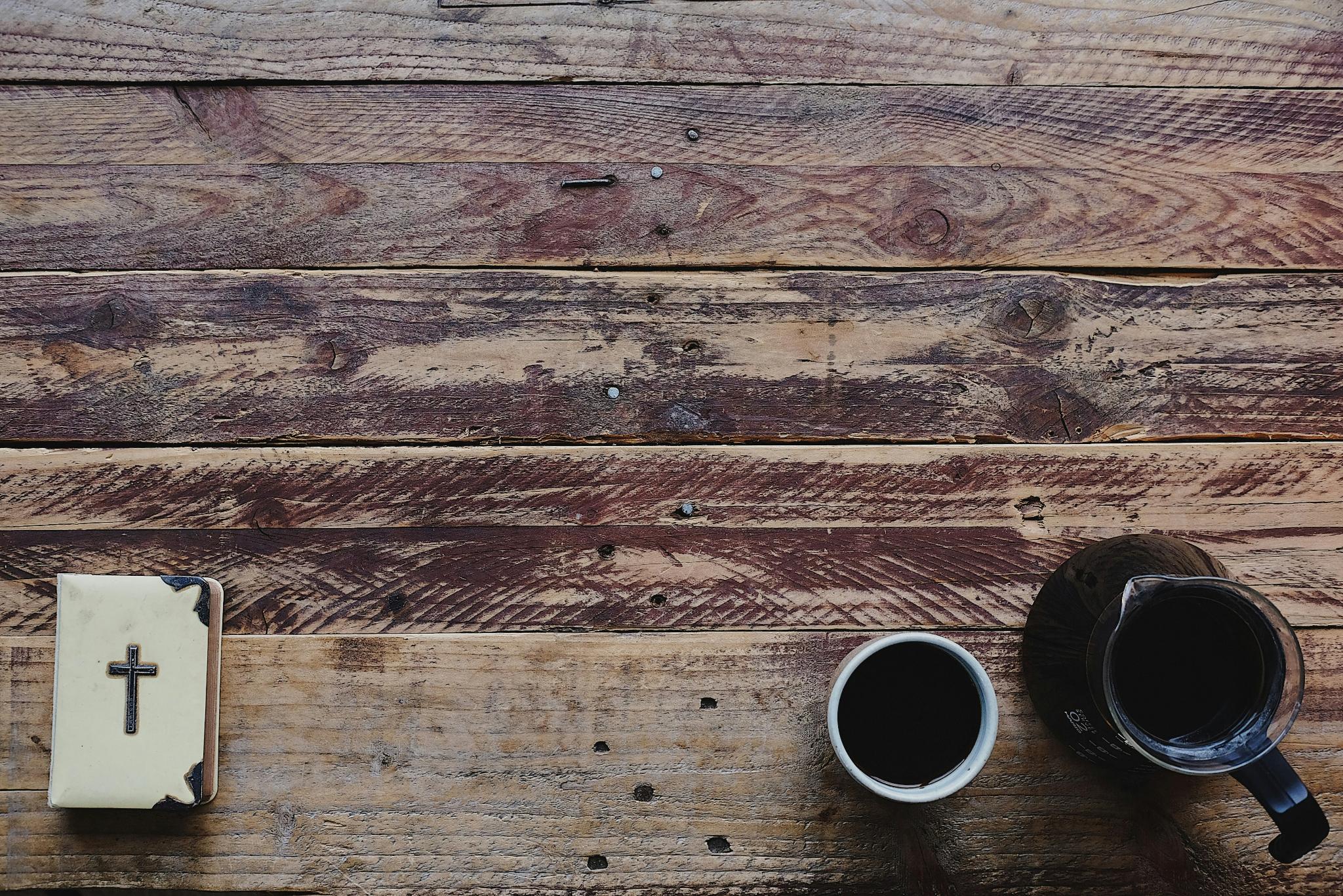 Auf einem alten Holztisch liegt links unten eine weiße Bibel mit einem Kreuz und Verzierungen aus Silber und au der rechten Seite eine Kanne Kaffee, daneben steht ein Becher mit Kaffee