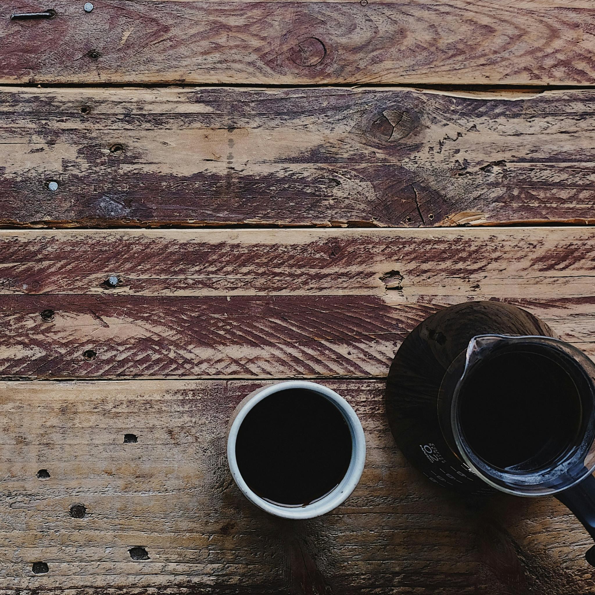 Auf einem alten Holztisch liegt links unten eine weiße Bibel mit einem Kreuz und Verzierungen aus Silber und au der rechten Seite eine Kanne Kaffee, daneben steht ein Becher mit Kaffee
