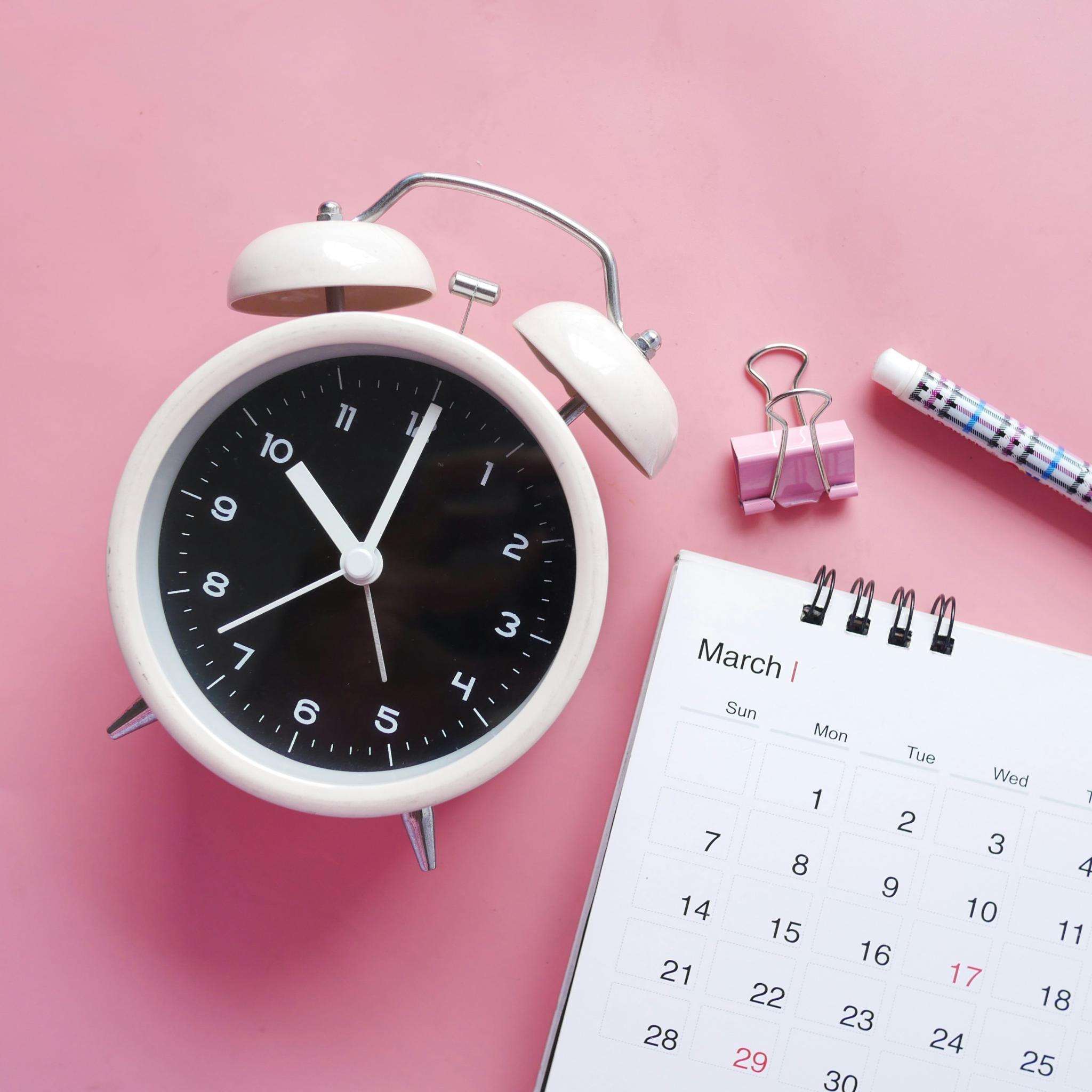 Blick von oben auf einen rosafarbenen Schreibtisch. Man sieht einen Wecker, einen Monatskalender, Stift und Heftklammer