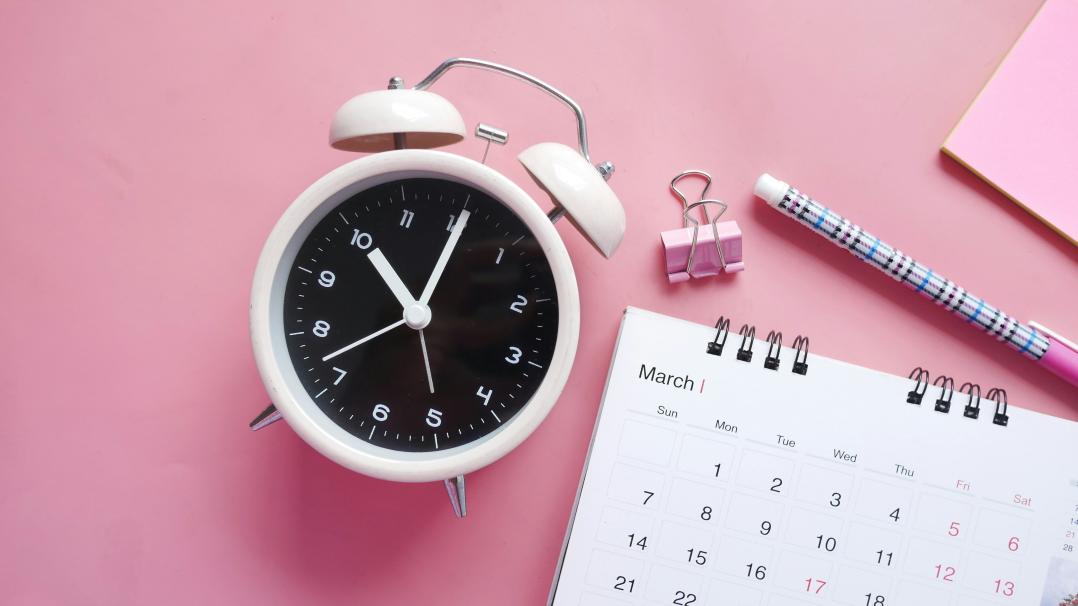 Blick von oben auf einen rosafarbenen Schreibtisch. Man sieht einen Wecker, einen Monatskalender, Stift und Heftklammer