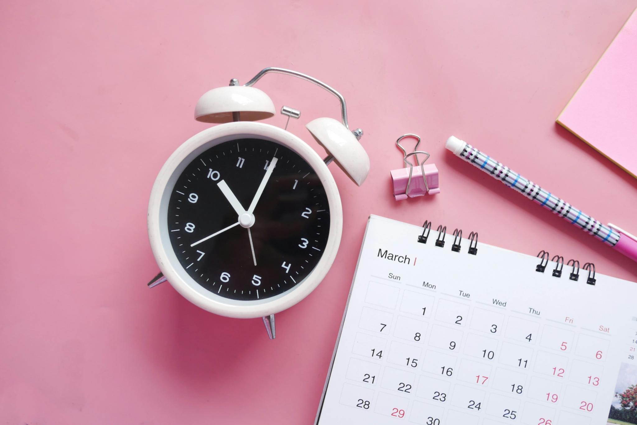 Blick von oben auf einen rosafarbenen Schreibtisch. Man sieht einen Wecker, einen Monatskalender, Stift und Heftklammer