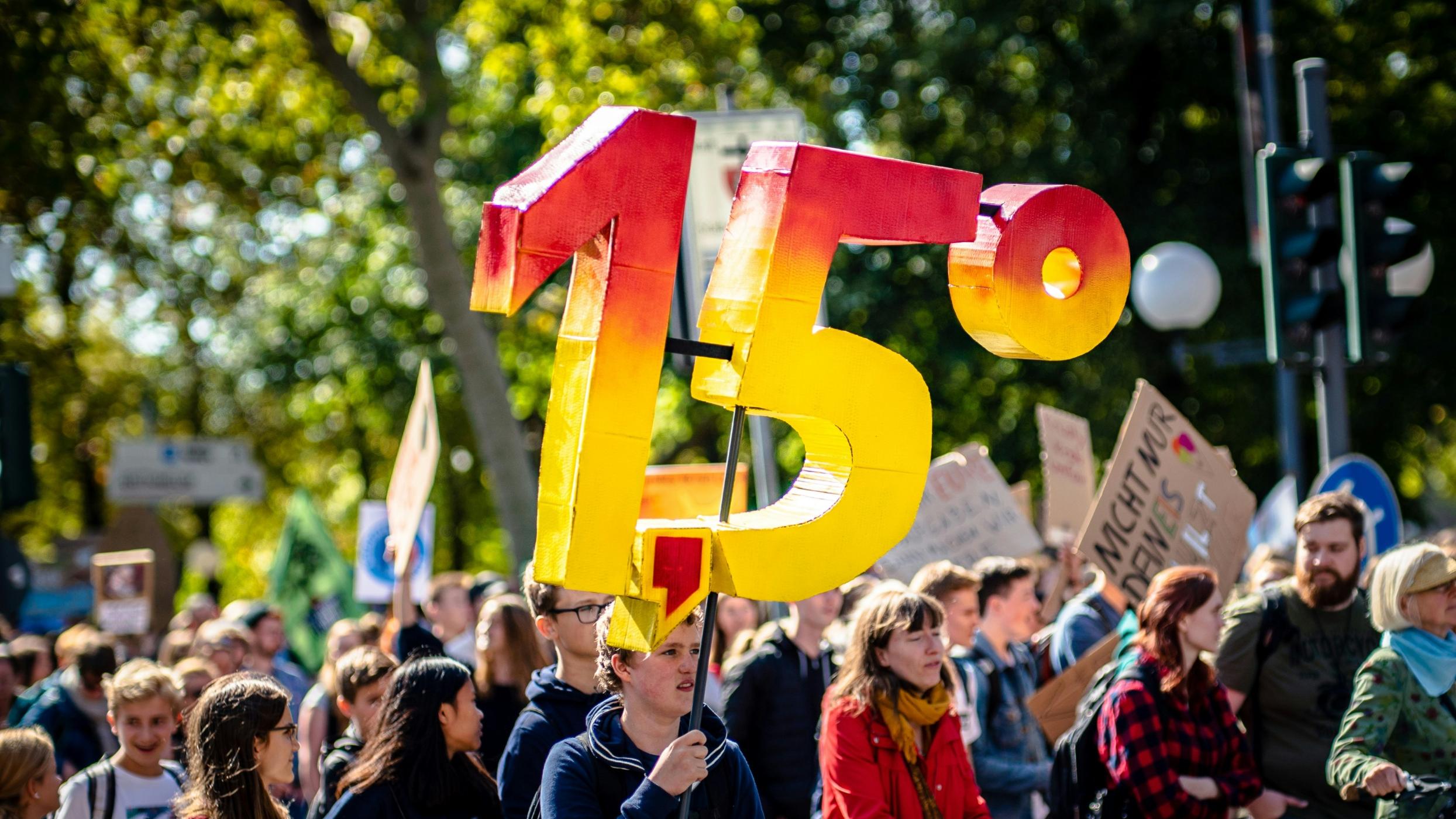 Man sieht eine Gruppe von Demonstrierenden mit Plakaten. Eine Person hält die Zahl '1,5 Grad' aus Pappe in die Luft