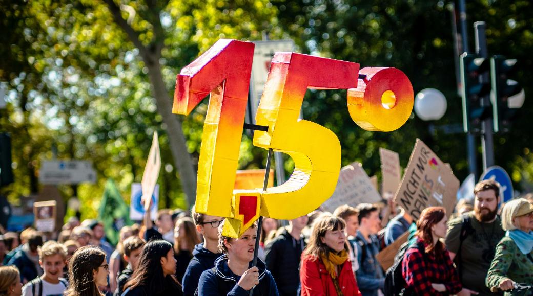 Man sieht eine Gruppe von Demonstrierenden mit Plakaten. Eine Person hält die Zahl '1,5 Grad' aus Pappe in die Luft