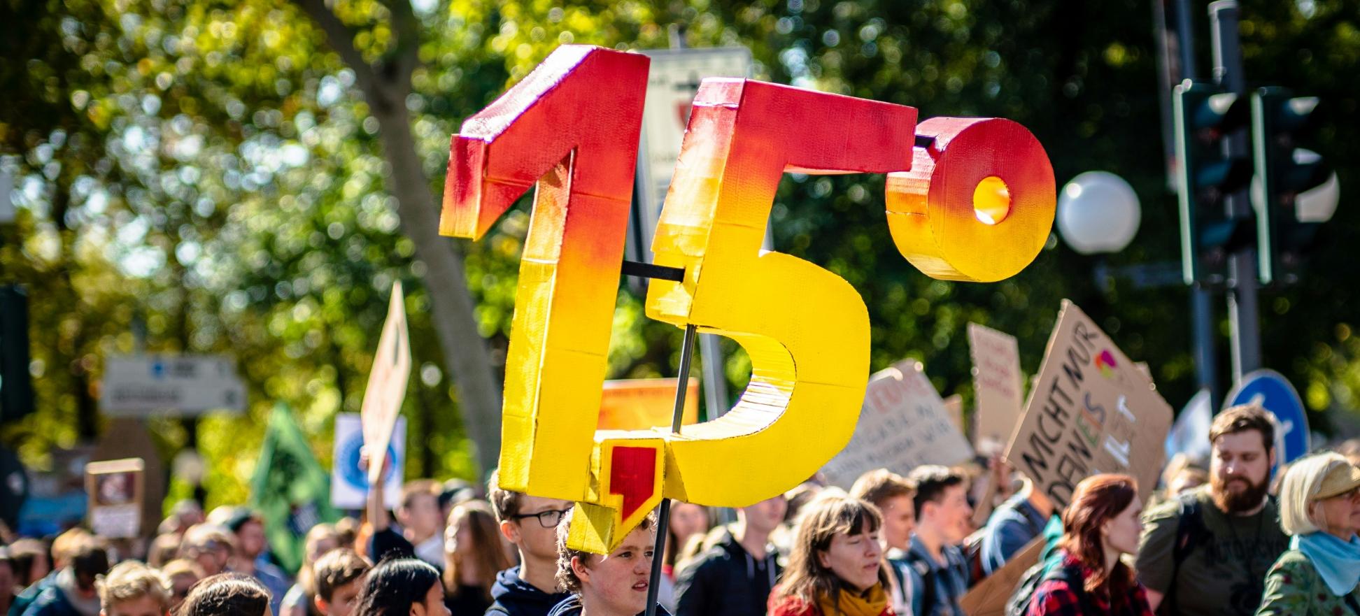 Man sieht eine Gruppe von Demonstrierenden mit Plakaten. Eine Person hält die Zahl '1,5 Grad' aus Pappe in die Luft