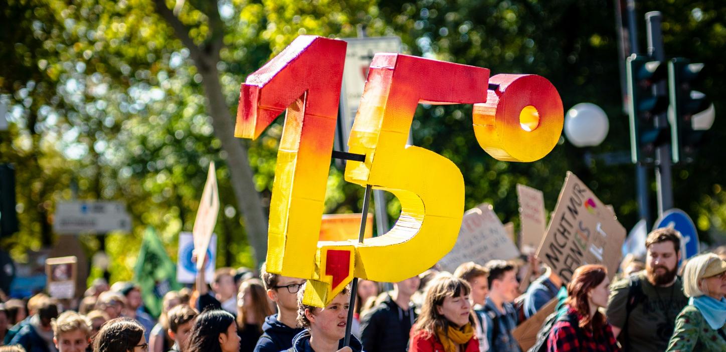 Man sieht eine Gruppe von Demonstrierenden mit Plakaten. Eine Person hält die Zahl '1,5 Grad' aus Pappe in die Luft