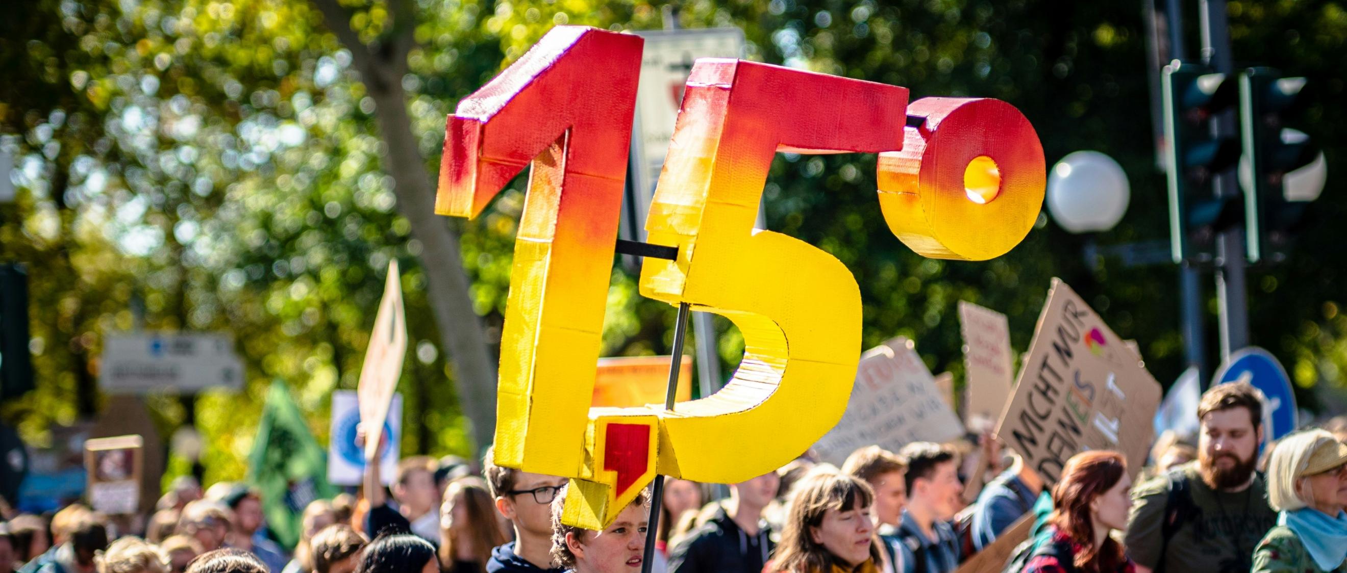 Man sieht eine Gruppe von Demonstrierenden mit Plakaten. Eine Person hält die Zahl '1,5 Grad' aus Pappe in die Luft