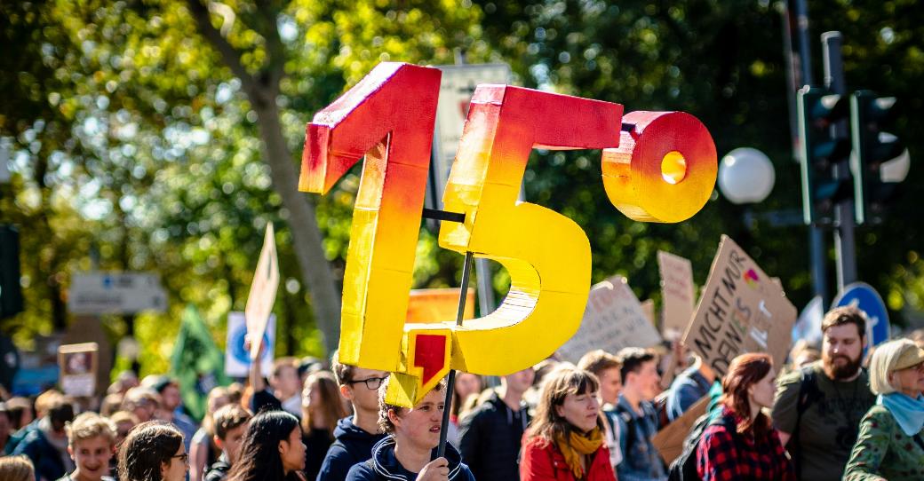 Man sieht eine Gruppe von Demonstrierenden mit Plakaten. Eine Person hält die Zahl '1,5 Grad' aus Pappe in die Luft