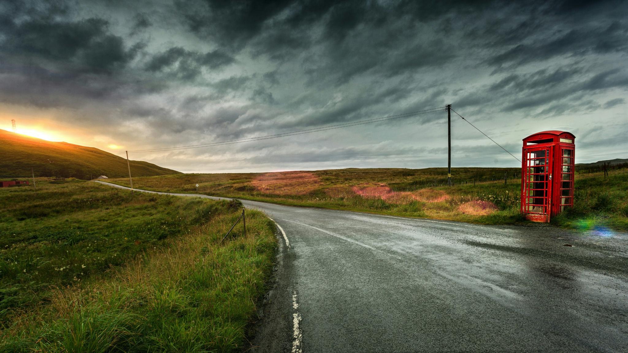 Eine einsame Straße in einer grünen Landschaft mit Sonnenuntergang, am Straßenrand steht eine alte englische rote Telefonzelle