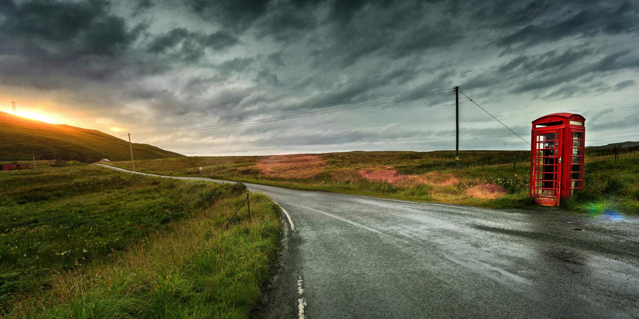 Eine einsame Straße in einer grünen Landschaft mit Sonnenuntergang, am Straßenrand steht eine alte englische rote Telefonzelle