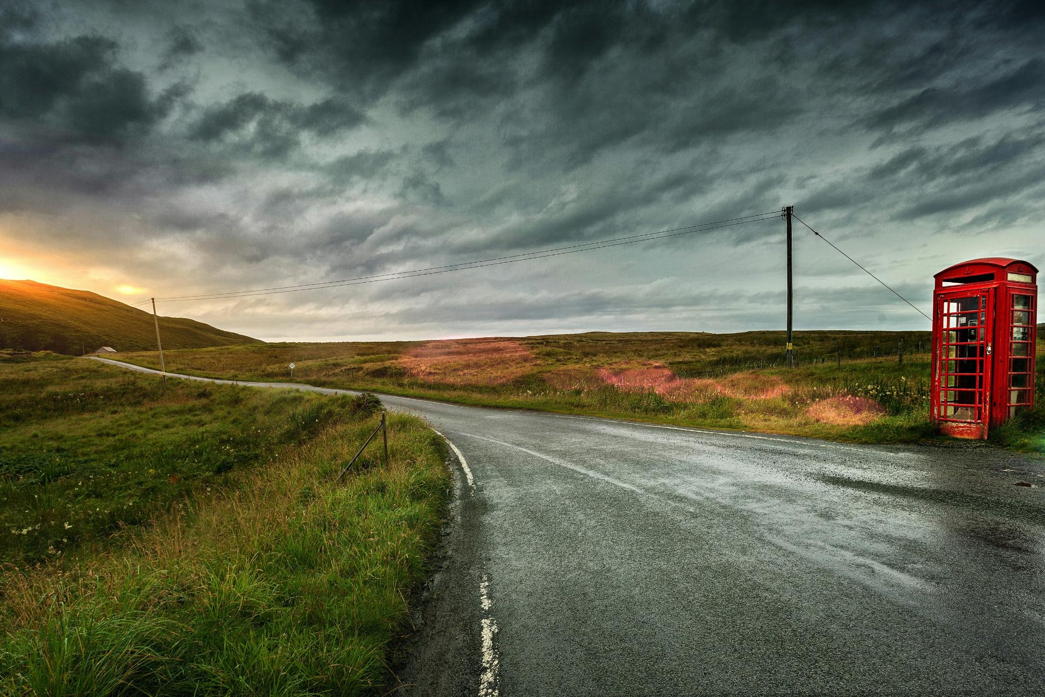 Eine einsame Straße in einer grünen Landschaft mit Sonnenuntergang, am Straßenrand steht eine alte englische rote Telefonzelle