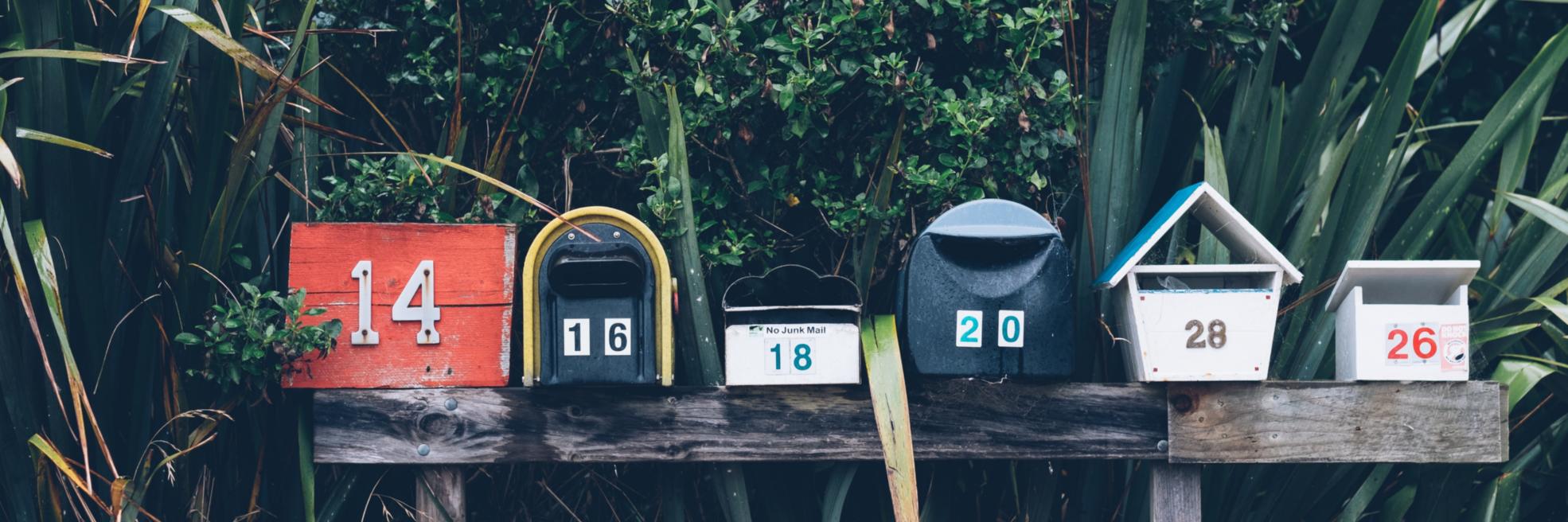 Man sieht mehrere bunte Briefkästen nebeneinander auf einem Balken