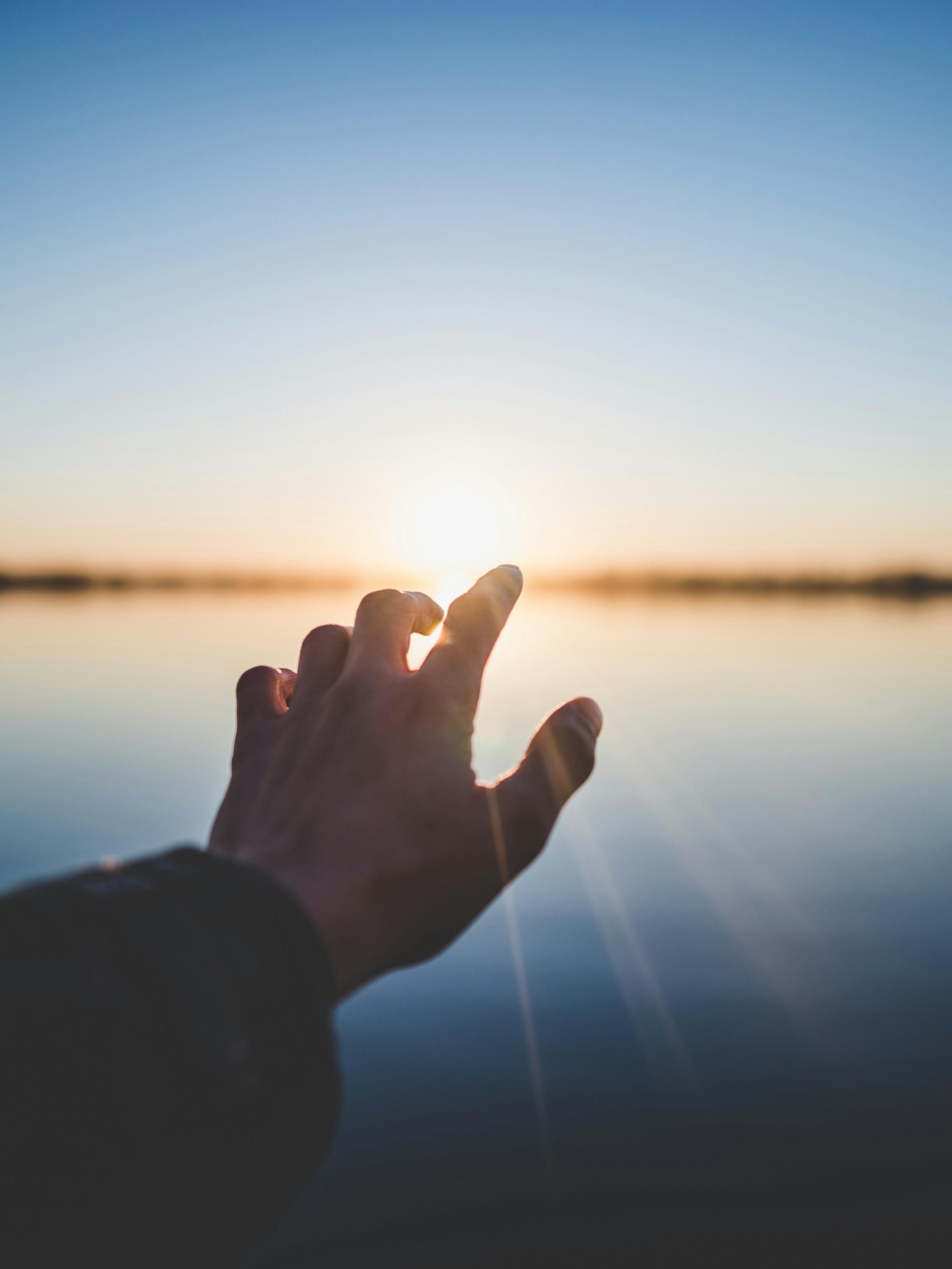 Eine Hand wird Richtung eines Gewässers ausgestreckt und im Hintergrund geht die Sonne unter