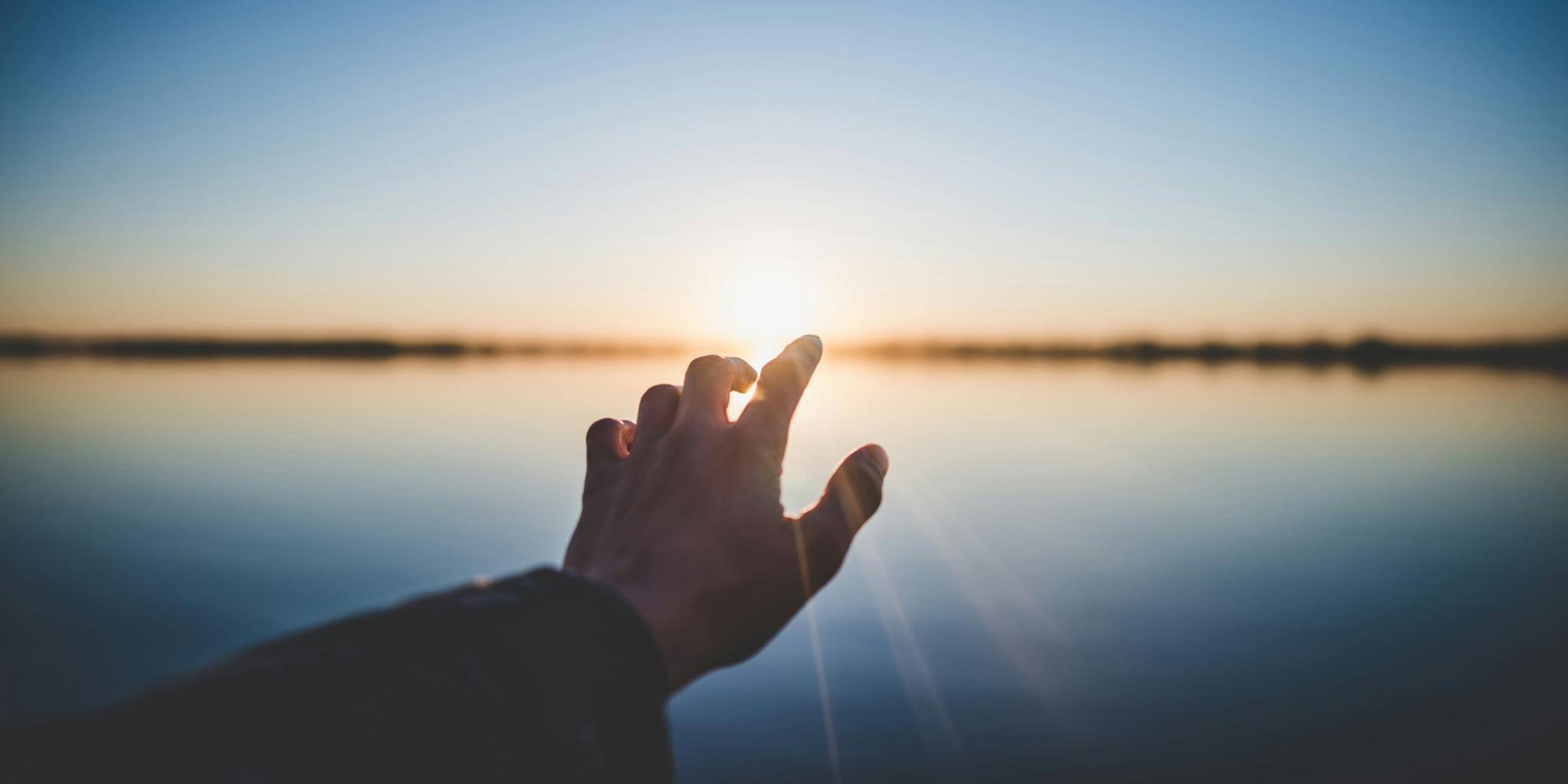 Eine Hand wird Richtung eines Gewässers ausgestreckt und im Hintergrund geht die Sonne unter