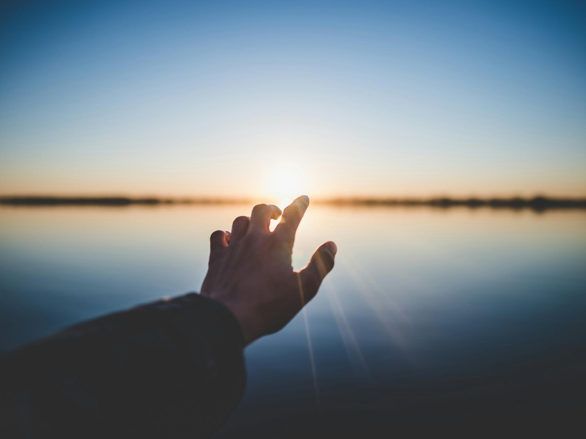 Eine Hand wird Richtung eines Gewässers ausgestreckt und im Hintergrund geht die Sonne unter