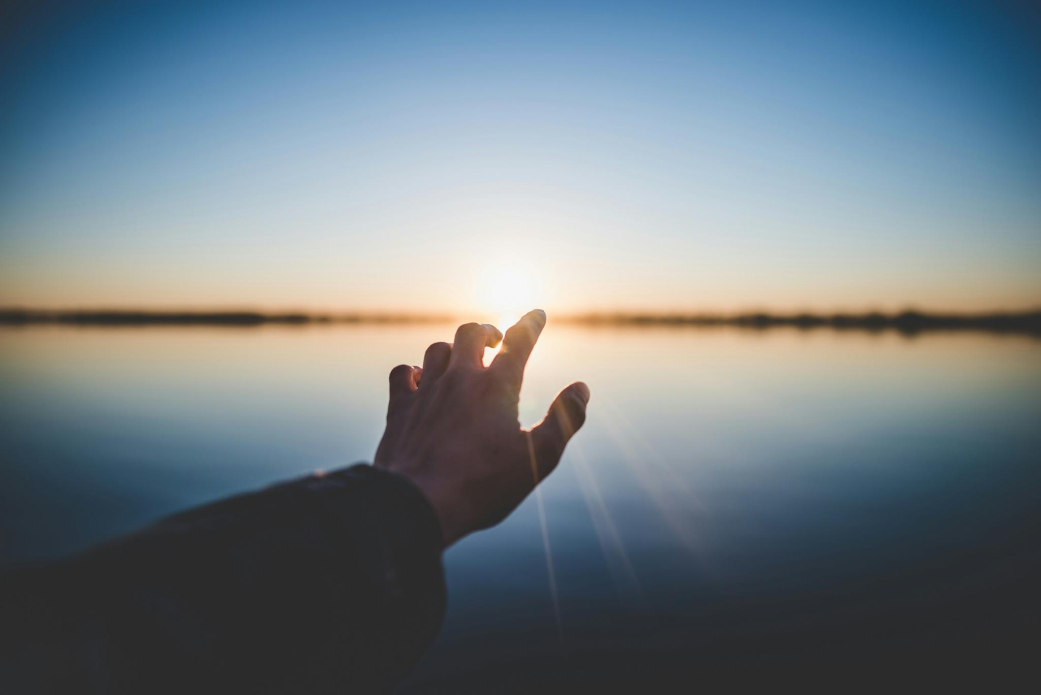 Eine Hand wird Richtung eines Gewässers ausgestreckt und im Hintergrund geht die Sonne unter