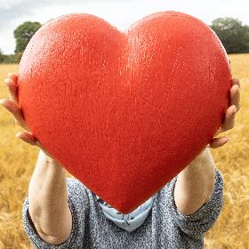Ein Person steht in einem trockenen Gräserfeld und hält ein großes rotes Herz in den Händen