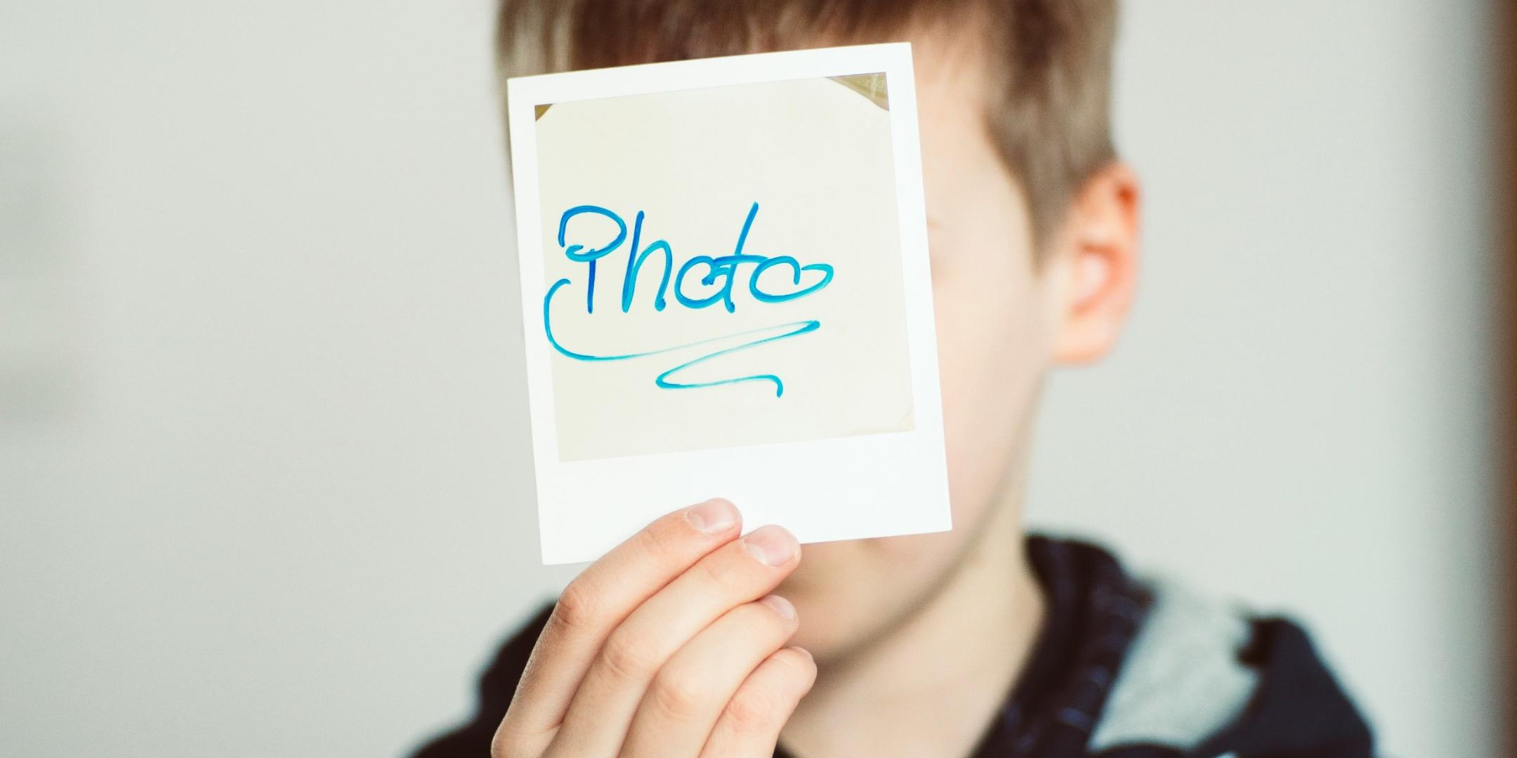 Man sieht einen Jungen, der ein Polaroid-Foto mit der Aufschrift 'Photo' in die Kamera hält