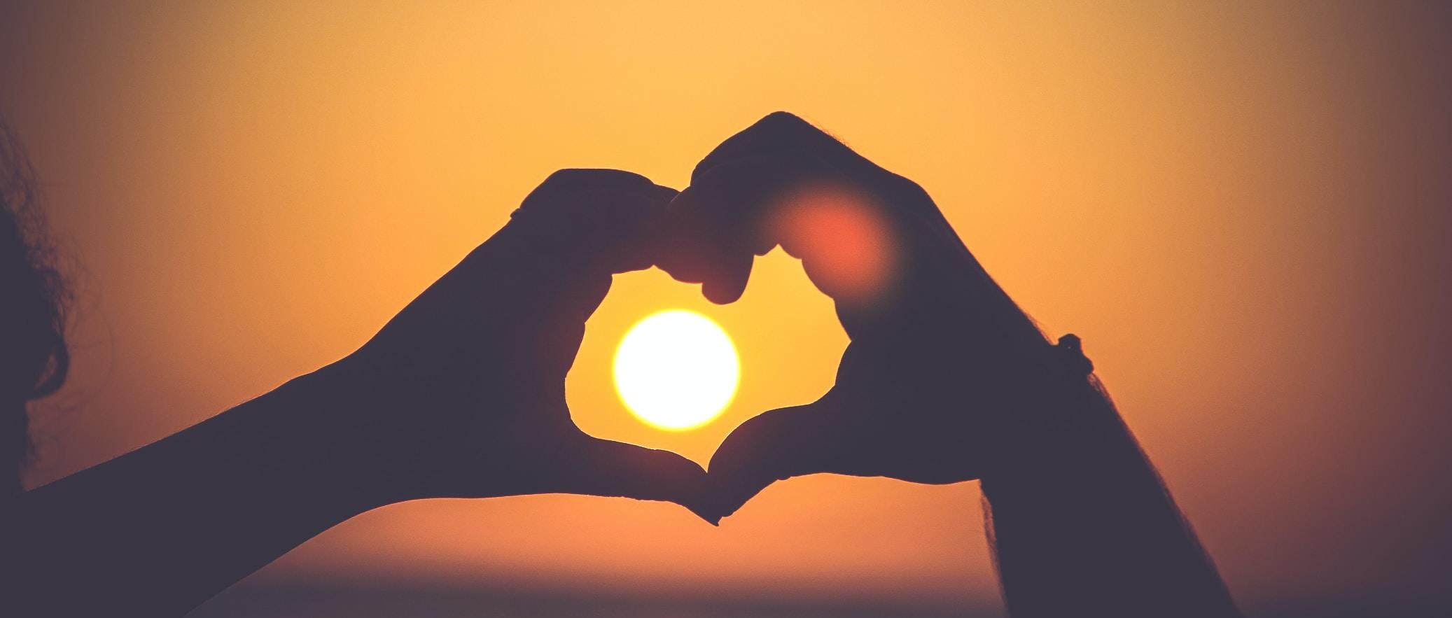 Zwei Hände formen ein Herz um die untergehende Sonne