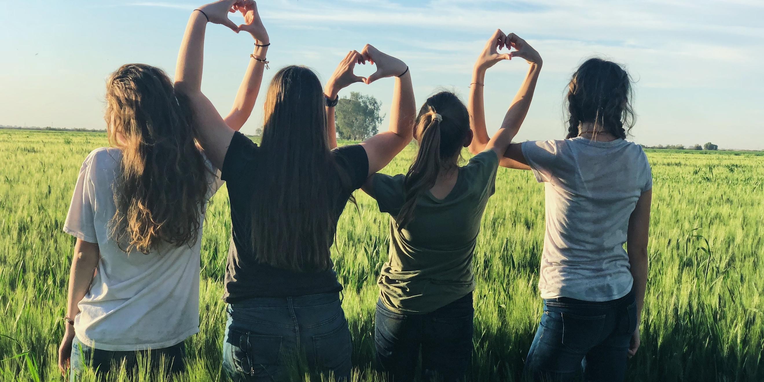 Vier junge Frauen stehen in einem Feld und formen ihre Hände zu Herzen und bilden damit eine Kette.