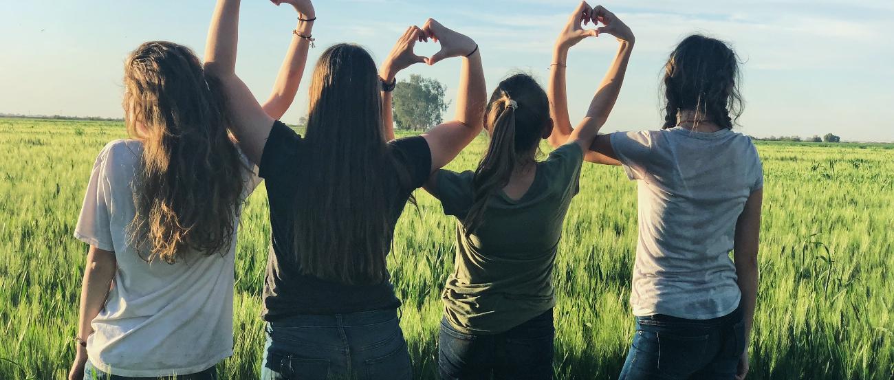 Vier junge Frauen stehen in einem Feld und formen ihre Hände zu Herzen und bilden damit eine Kette.