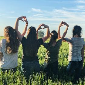 Vier junge Frauen stehen in einem Feld und formen ihre Hände zu Herzen und bilden damit eine Kette.