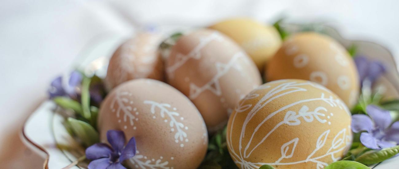 Auf einem Teller liegen braune Eiern, die mit weißen Verzierungen bemalt wurden. Dekoriert wurden sie auf dem Teller mit grünen Blättern und Blüten in violett