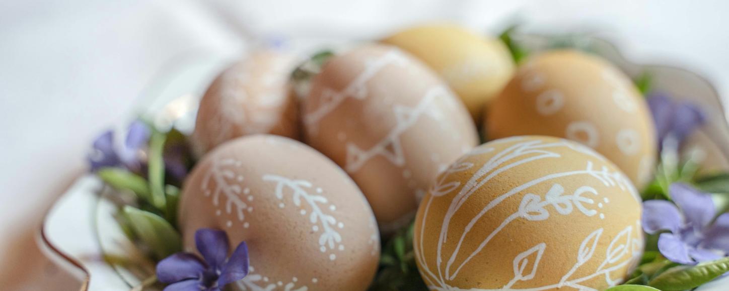 Auf einem Teller liegen braune Eiern, die mit weißen Verzierungen bemalt wurden. Dekoriert wurden sie auf dem Teller mit grünen Blättern und Blüten in violett