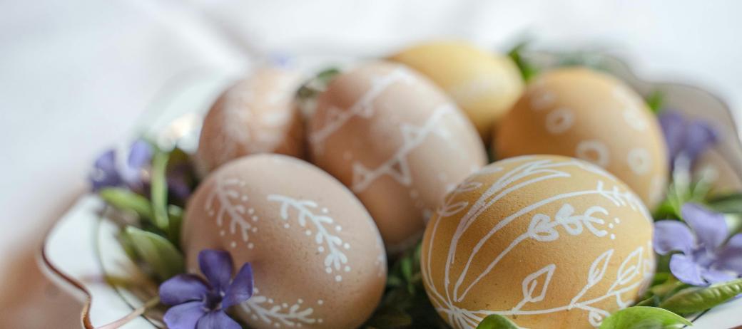 Auf einem Teller liegen braune Eiern, die mit weißen Verzierungen bemalt wurden. Dekoriert wurden sie auf dem Teller mit grünen Blättern und Blüten in violett