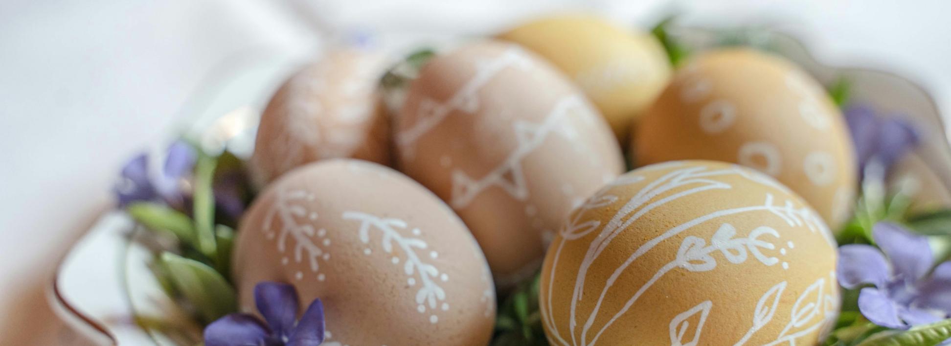 Auf einem Teller liegen braune Eiern, die mit weißen Verzierungen bemalt wurden. Dekoriert wurden sie auf dem Teller mit grünen Blättern und Blüten in violett