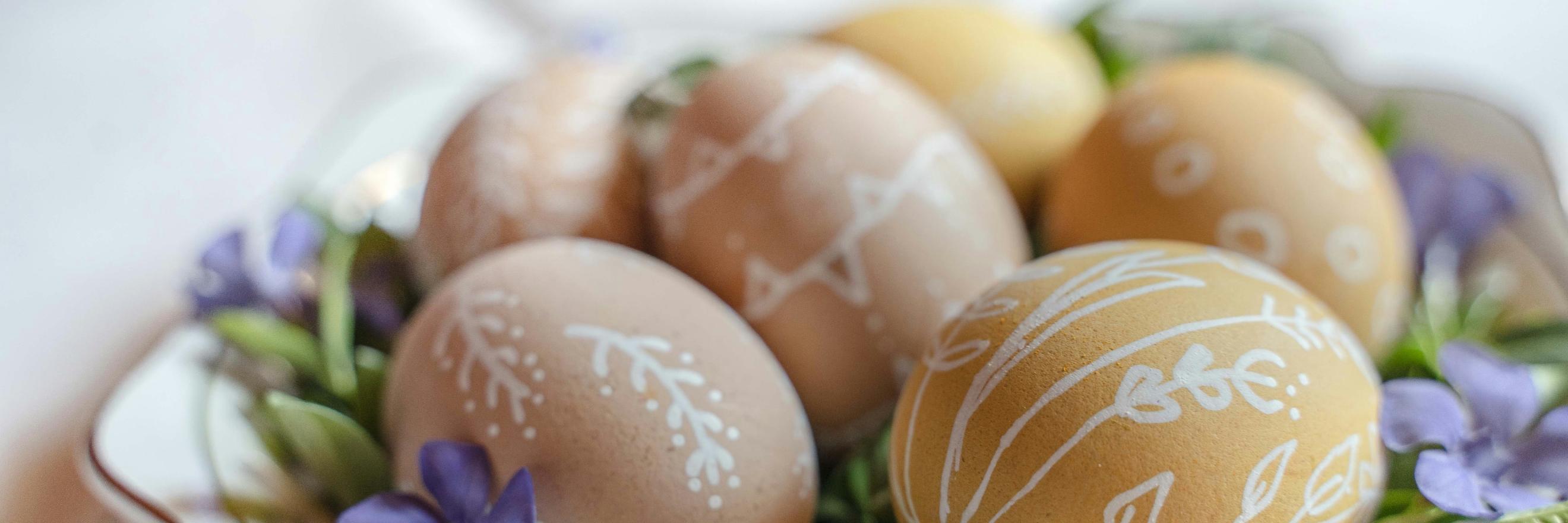 Auf einem Teller liegen braune Eiern, die mit weißen Verzierungen bemalt wurden. Dekoriert wurden sie auf dem Teller mit grünen Blättern und Blüten in violett