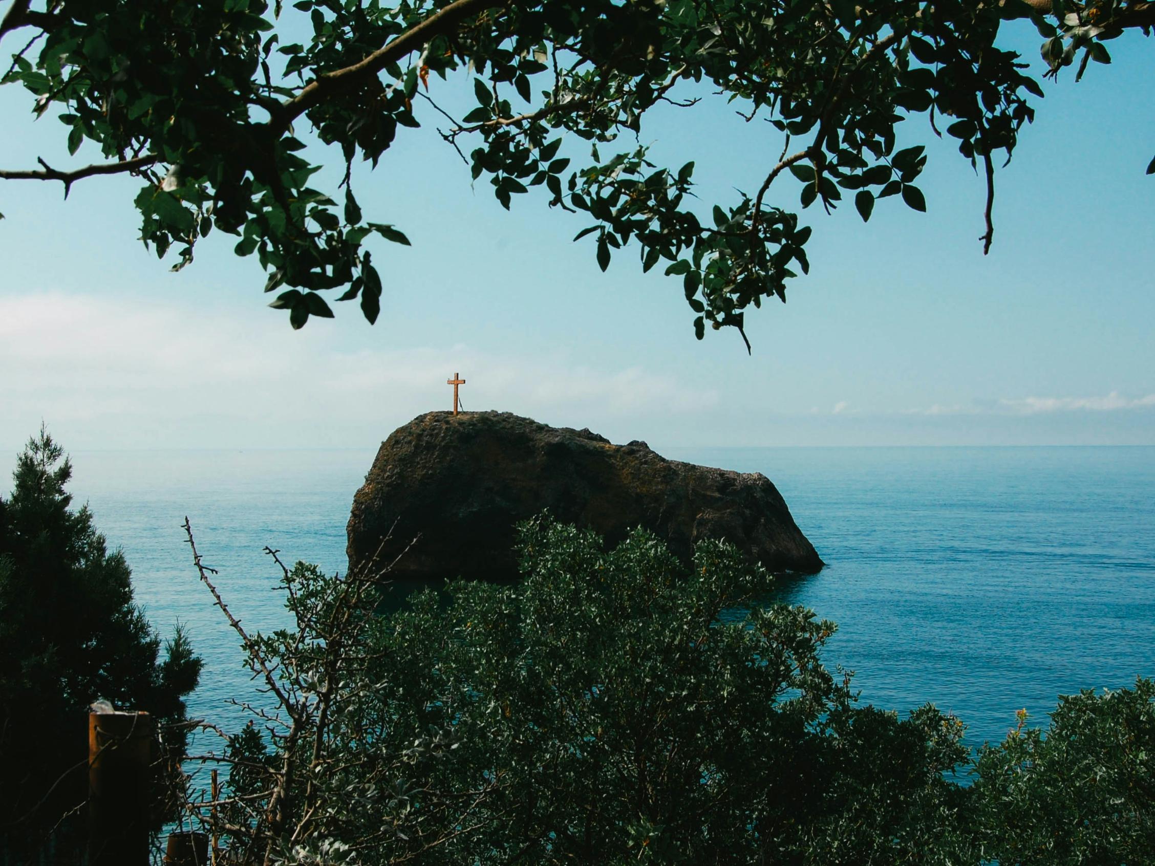 Man blickt vom Wald auf einen Felsen im Meer. Darauf steht ein großes Kreuz