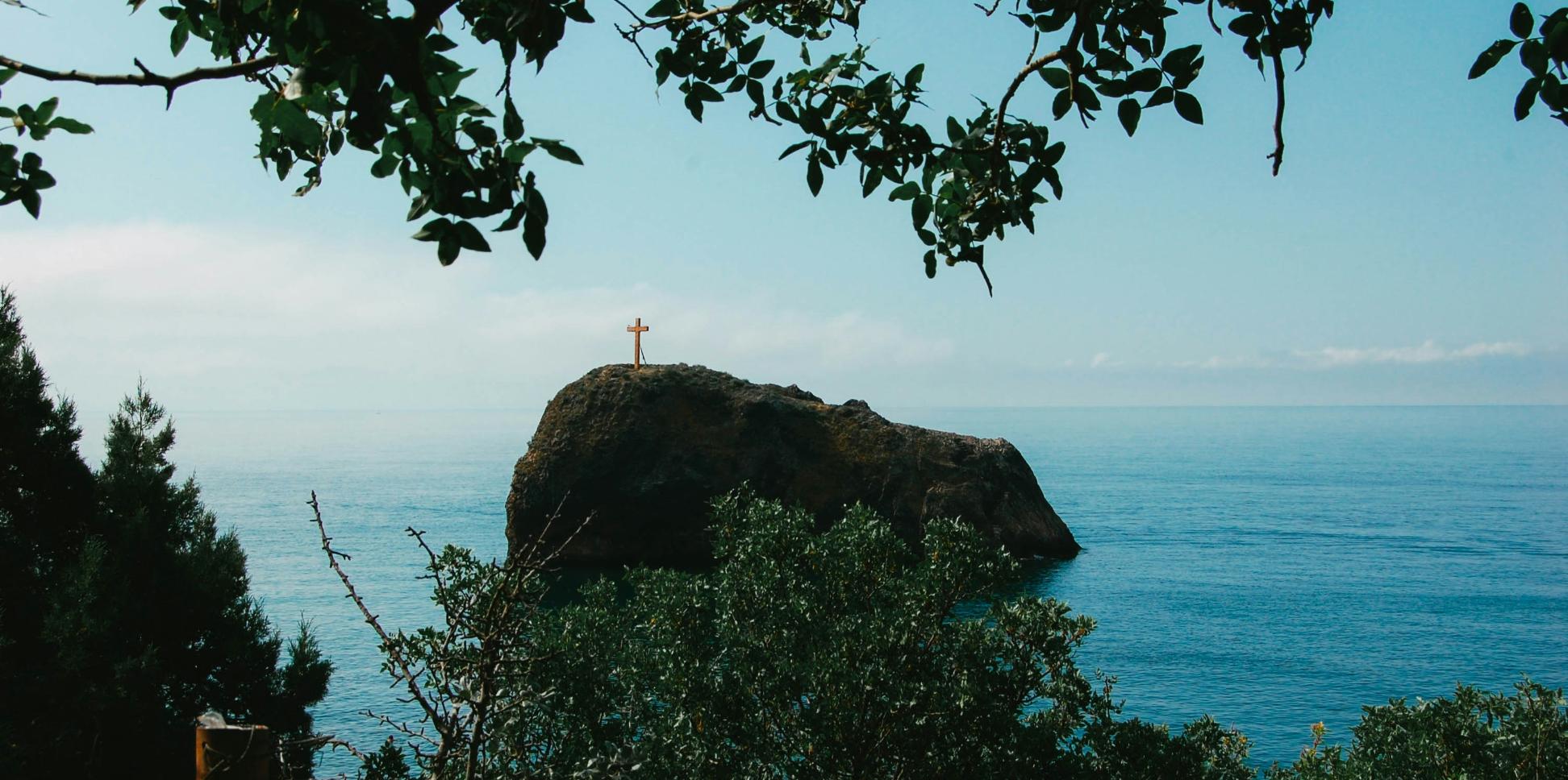Man blickt vom Wald auf einen Felsen im Meer. Darauf steht ein großes Kreuz