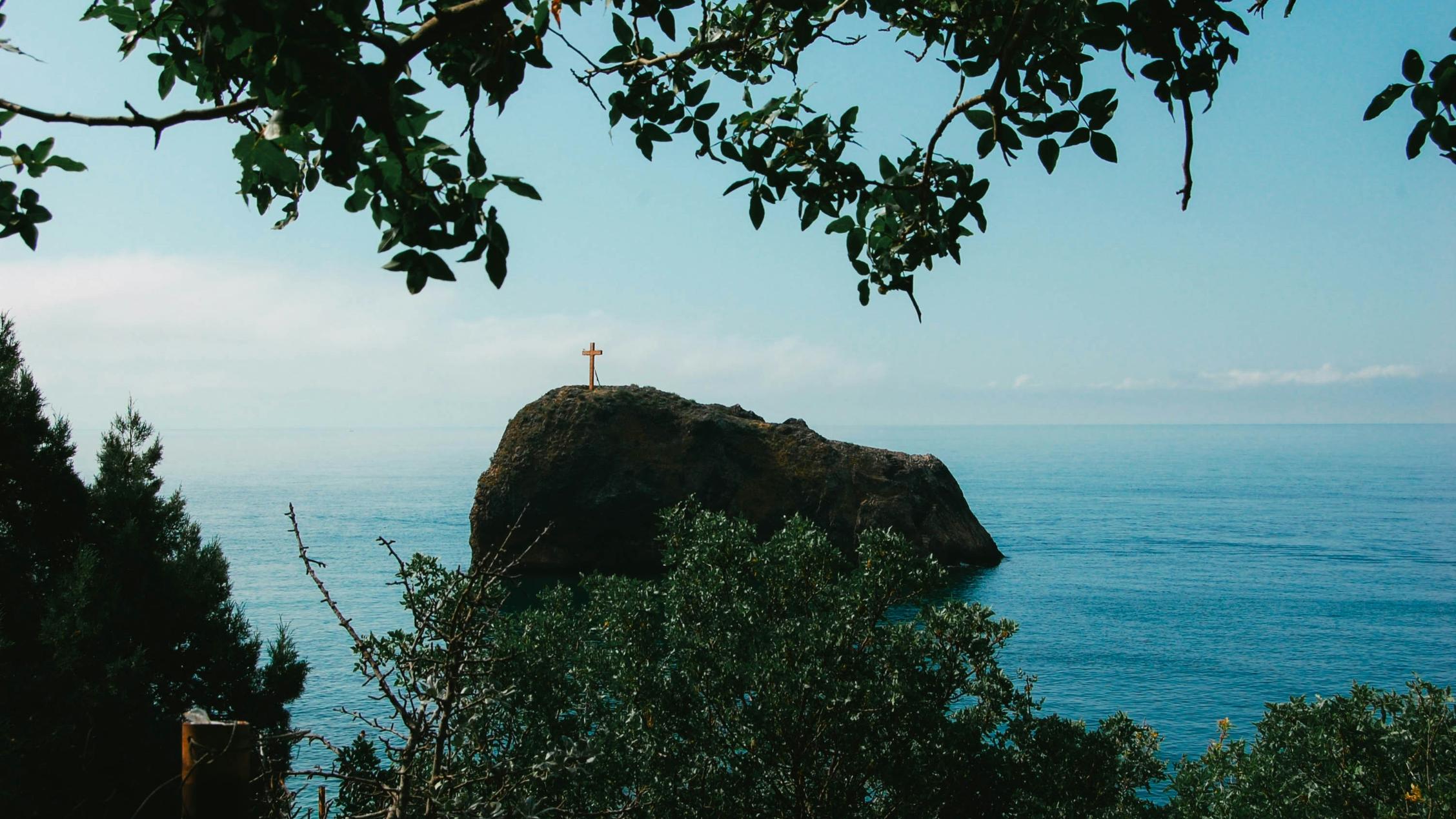 Man blickt vom Wald auf einen Felsen im Meer. Darauf steht ein großes Kreuz