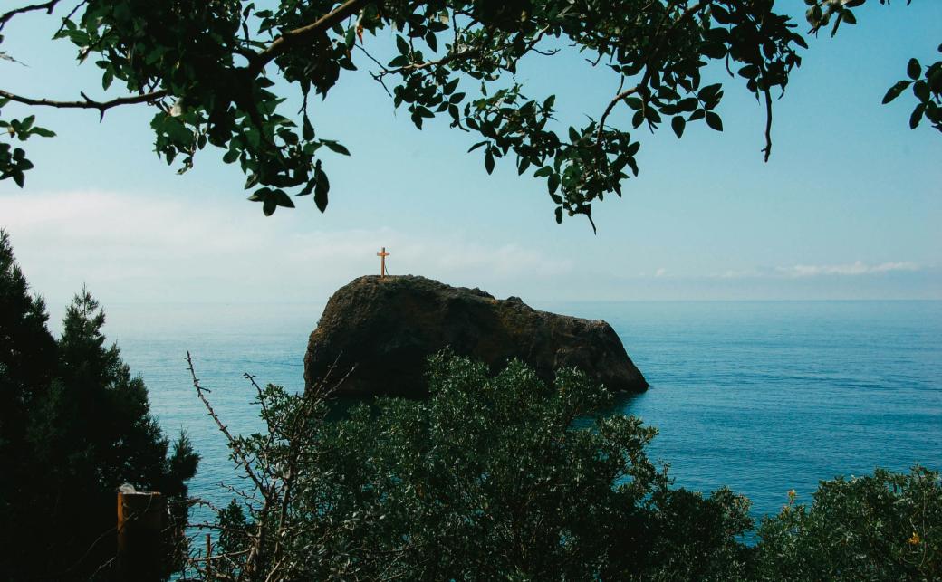 Man blickt vom Wald auf einen Felsen im Meer. Darauf steht ein großes Kreuz