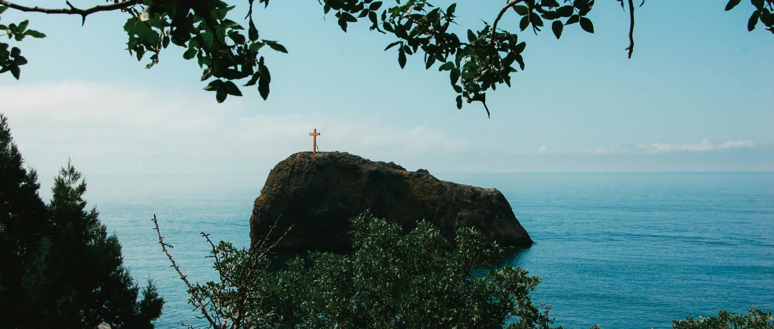 Man blickt vom Wald auf einen Felsen im Meer. Darauf steht ein großes Kreuz