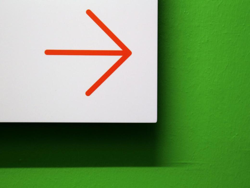 Vor einer grünen Wand ist ein weißes Hinweisschild mit einem roten Pfeil, der zur Seite zeigt