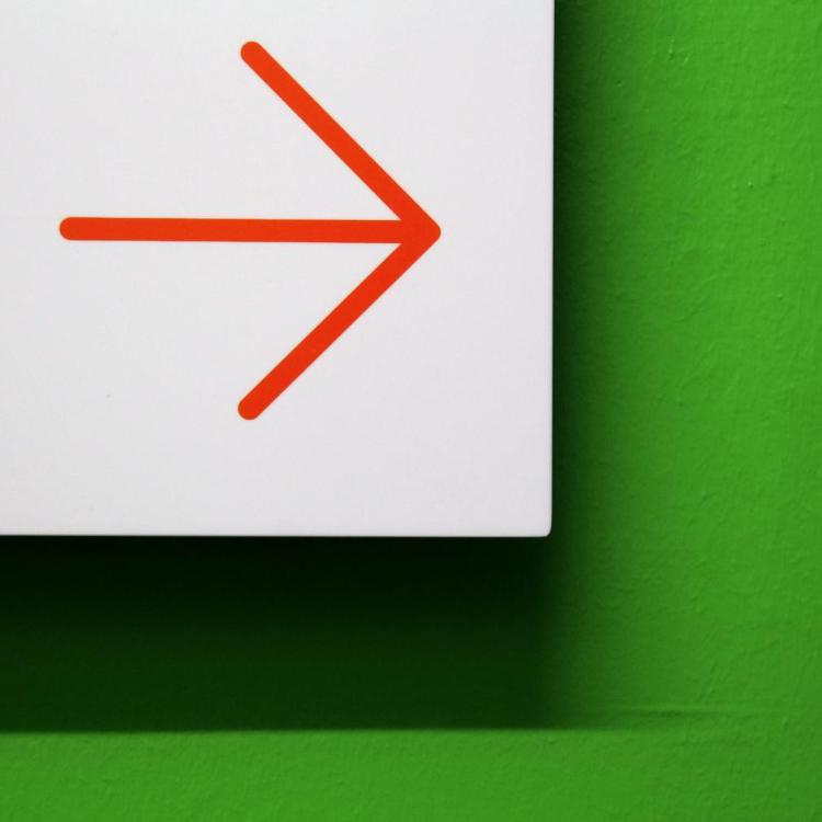 Vor einer grünen Wand ist ein weißes Hinweisschild mit einem roten Pfeil, der zur Seite zeigt