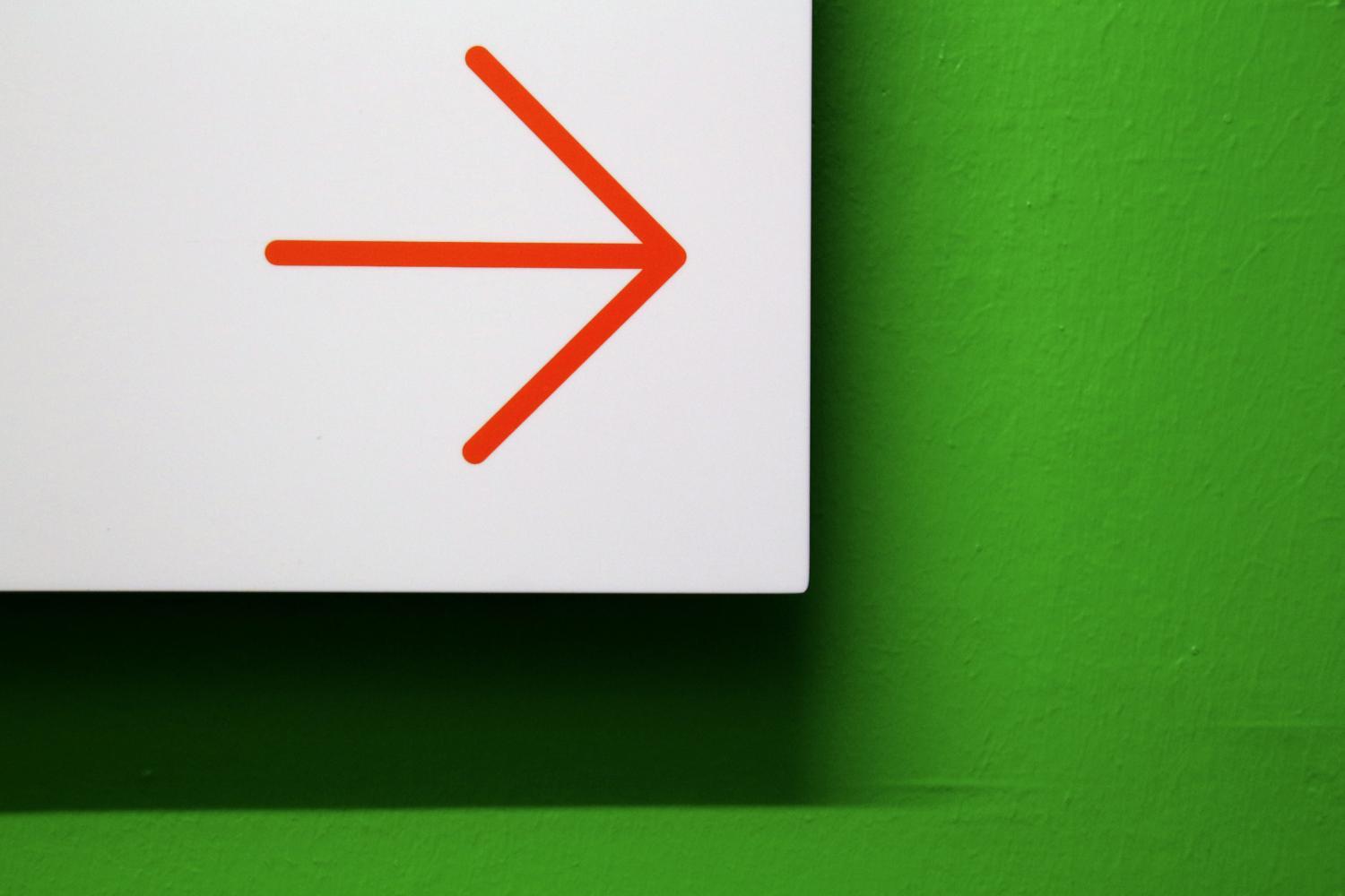 Vor einer grünen Wand ist ein weißes Hinweisschild mit einem roten Pfeil, der zur Seite zeigt