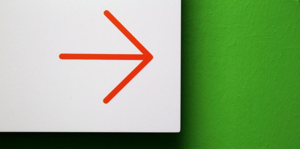 Vor einer grünen Wand ist ein weißes Hinweisschild mit einem roten Pfeil, der zur Seite zeigt