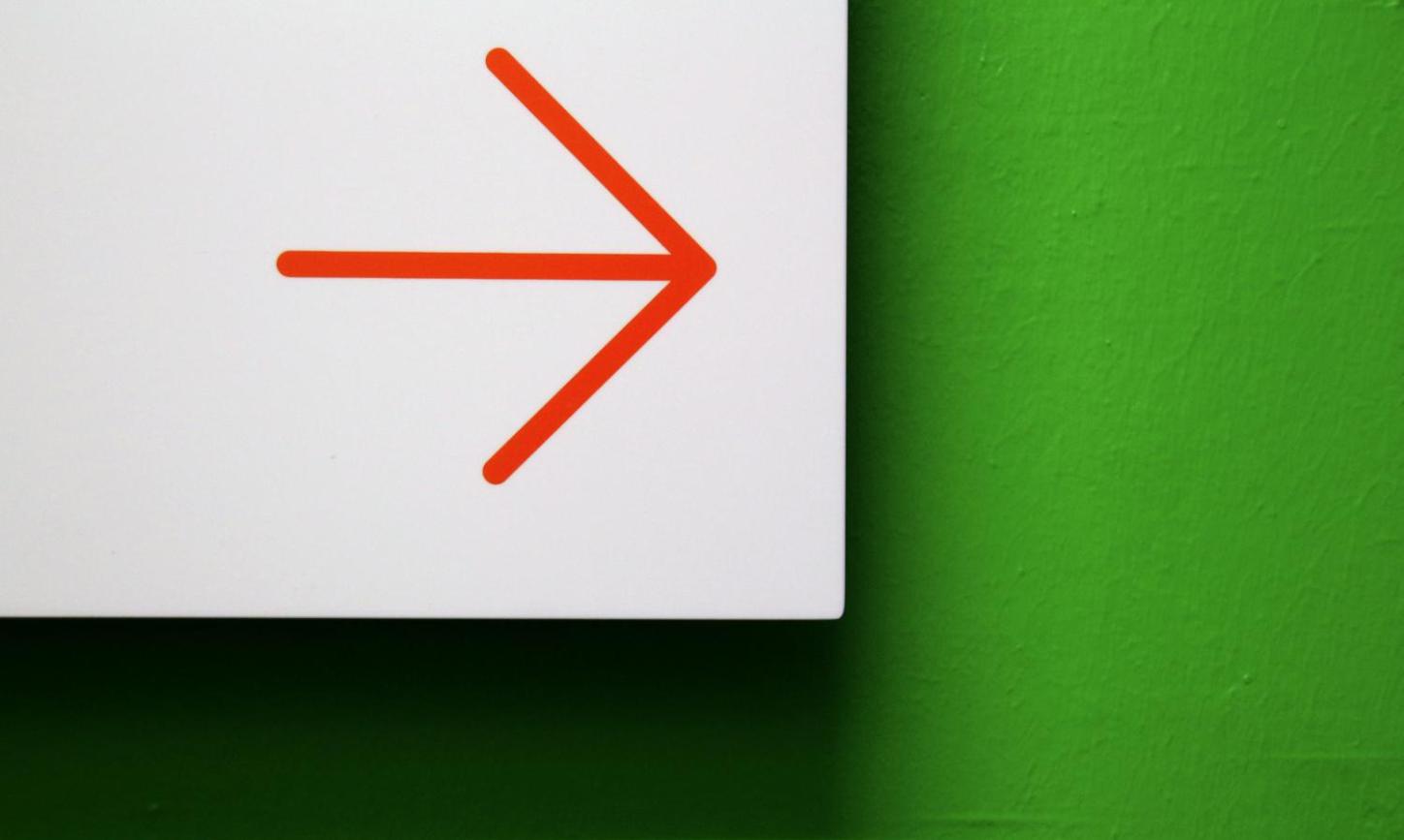 Vor einer grünen Wand ist ein weißes Hinweisschild mit einem roten Pfeil, der zur Seite zeigt