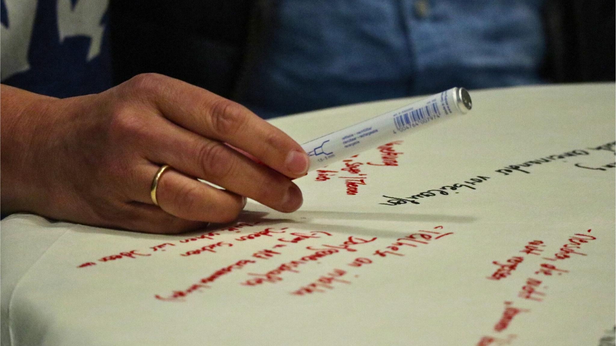 Ein Flipchartblatt liegt auf einem Tisch, es ist beschrieben. Eine Hand mit Stift schwebt darüber.