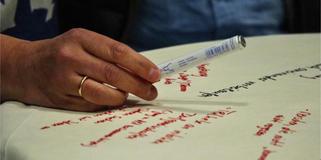 Ein Flipchartblatt liegt auf einem Tisch, es ist beschrieben. Eine Hand mit Stift schwebt darüber.