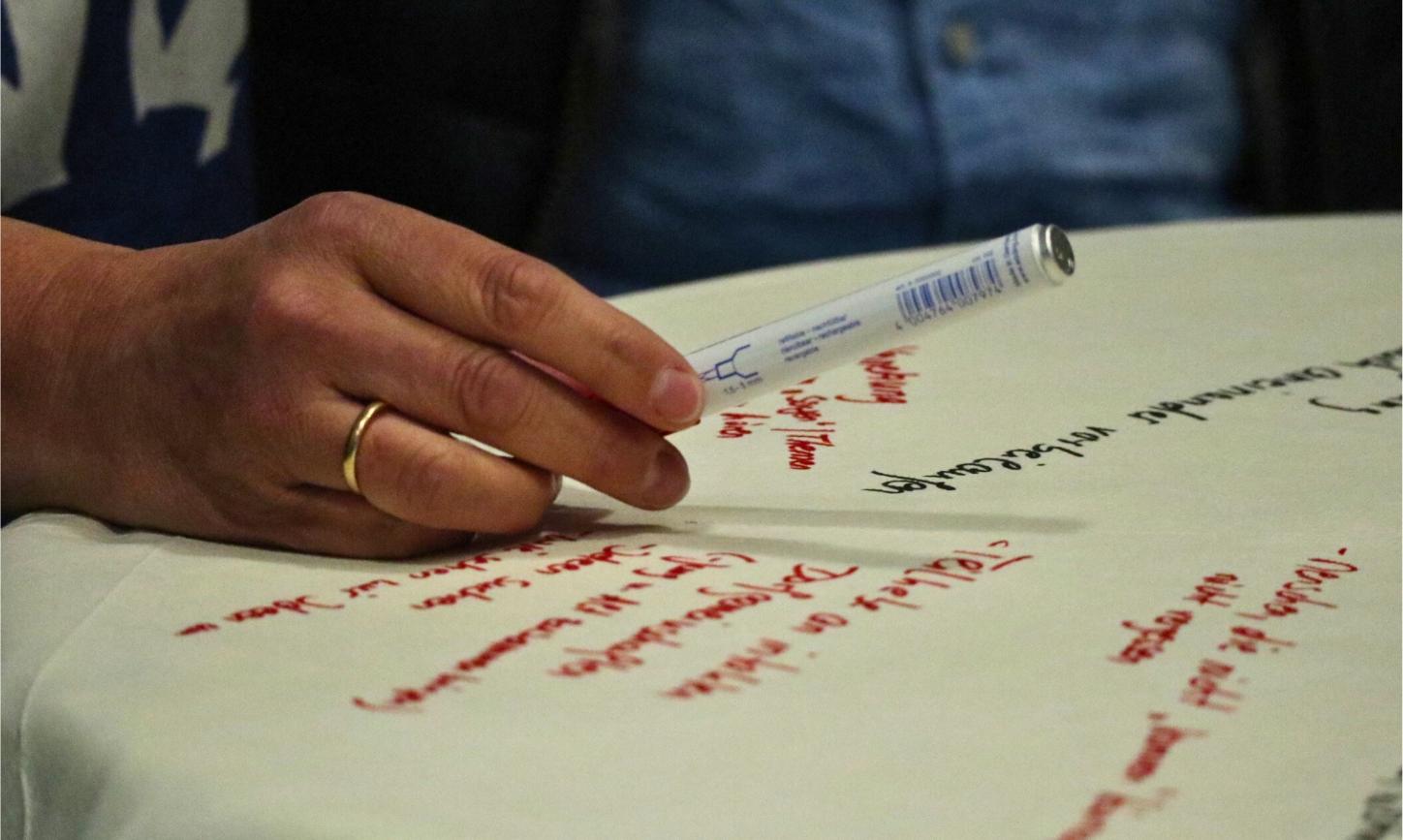 Ein Flipchartblatt liegt auf einem Tisch, es ist beschrieben. Eine Hand mit Stift schwebt darüber.