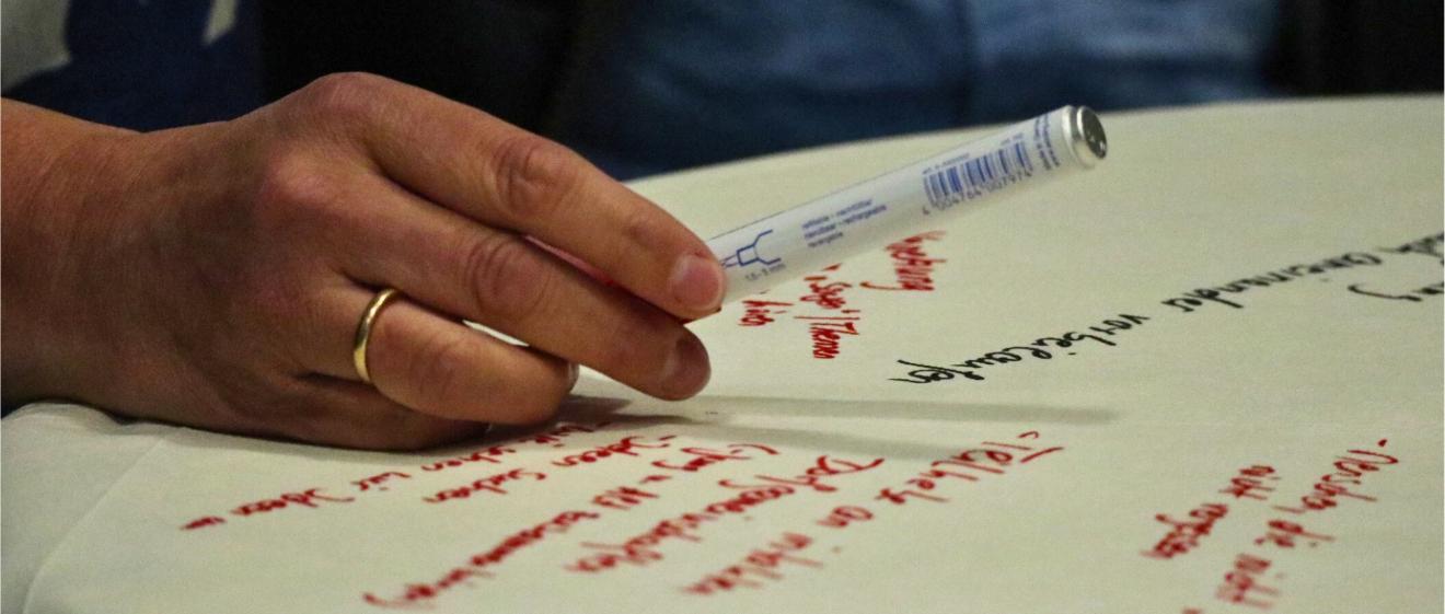 Ein Flipchartblatt liegt auf einem Tisch, es ist beschrieben. Eine Hand mit Stift schwebt darüber.