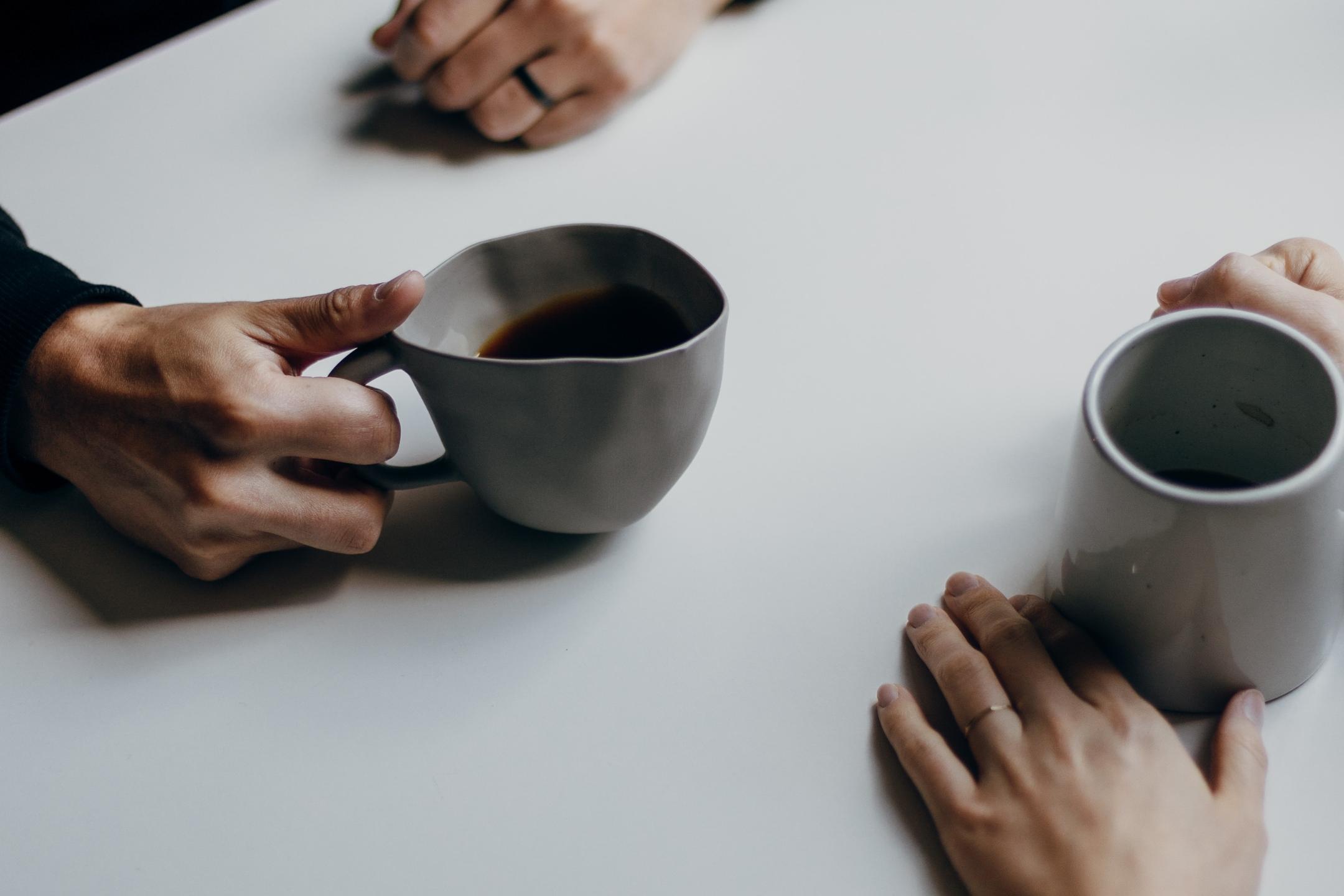 Auf einem Tisch stehen zwei Tassen Kaffee, daneben sind die Hände der Personen zu sehen, die am Tisch sitzen.