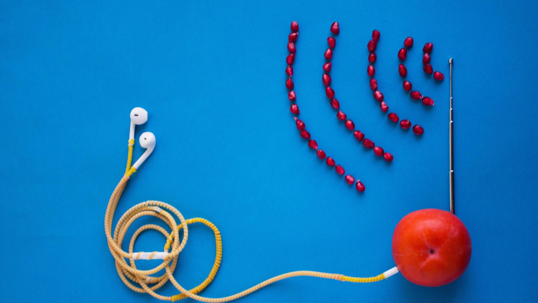 auf einer blauen Fläche liegt eine Mandarine, in der eine Antenne steckt. Vor der Antenne liegen  (in Form von Radiowellen) Granatapfelkerne. Von der Mandarine führt ein Kabel mit Kopfhöhrern, aus Nudeln.